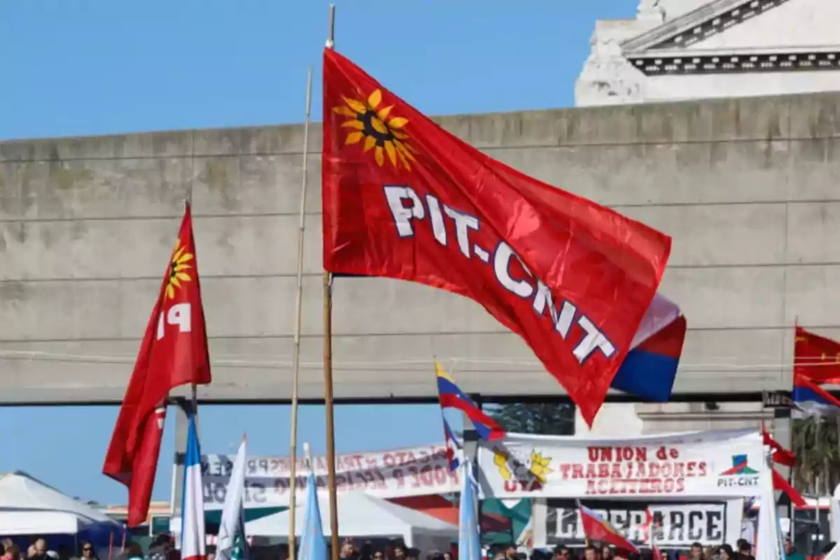 Bandera roja del PIT-CNT ondeando en una manifestación con otras banderas y pancartas al fondo