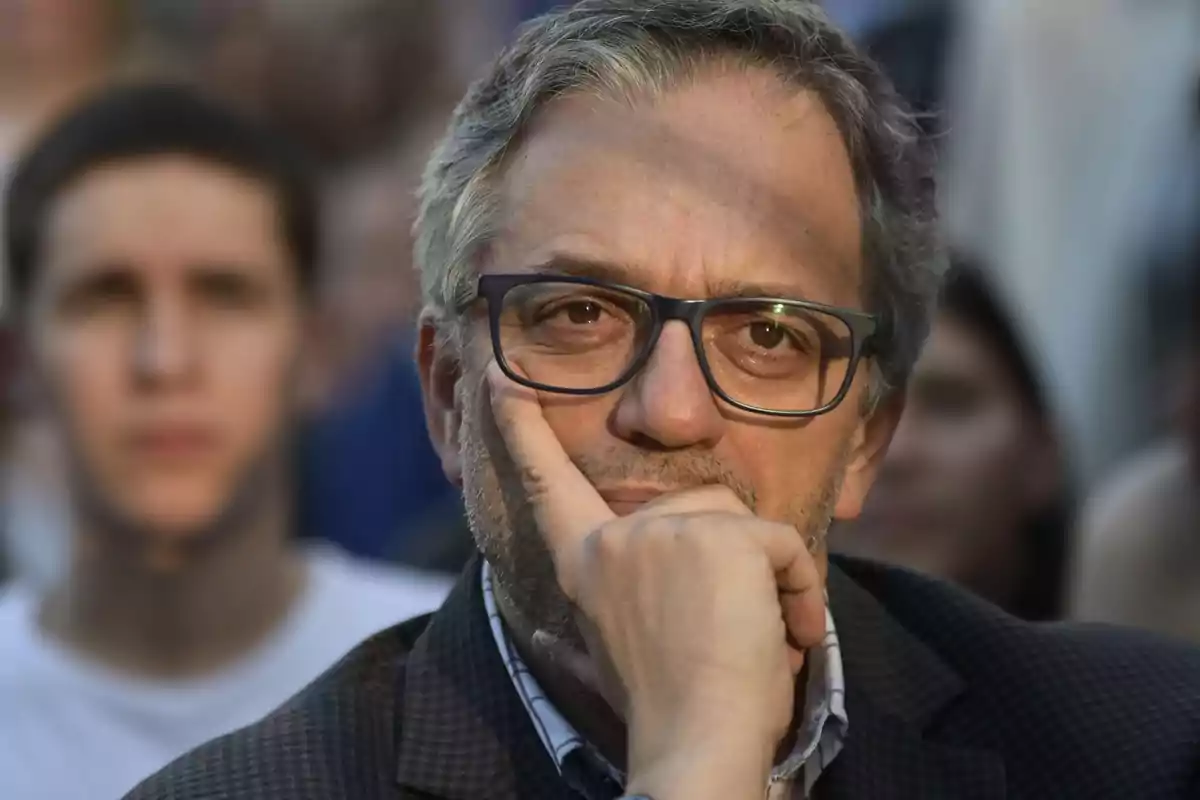 Hombre de mediana edad con gafas y barba corta mirando pensativo hacia el frente con la mano en el rostro y personas desenfocadas al fondo Hombre de mediana edad con gafas y barba corta mirando pensativo hacia el frente con la mano en el rostro y personas desenfocadas al fondo