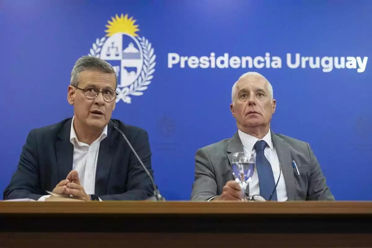 Dos hombres con traje y corbata sentados en una mesa durante una conferencia de prensa frente a un fondo azul con el escudo nacional y el texto Presidencia Uruguay