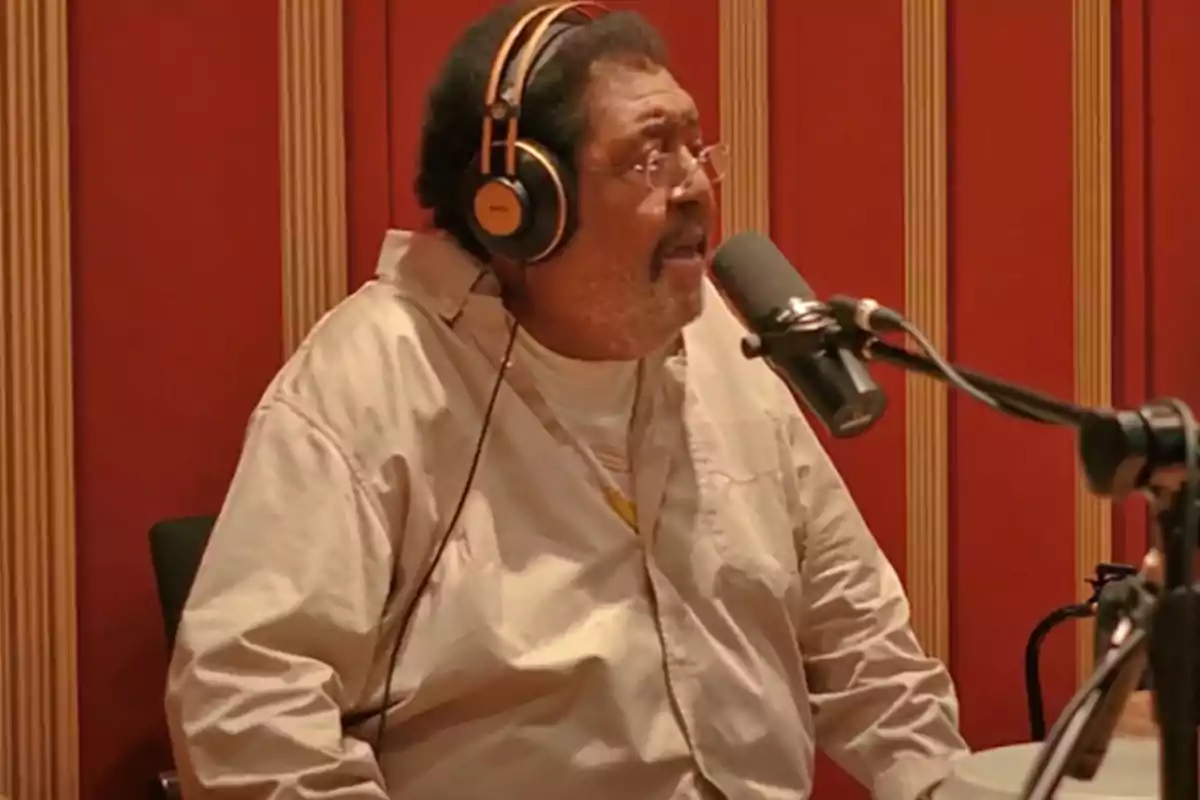 Hombre con audífonos y camisa beige hablando frente a un micrófono en un estudio de grabación con paredes rojas. Hombre con audífonos y camisa beige hablando frente a un micrófono en un estudio de grabación con paredes rojas.