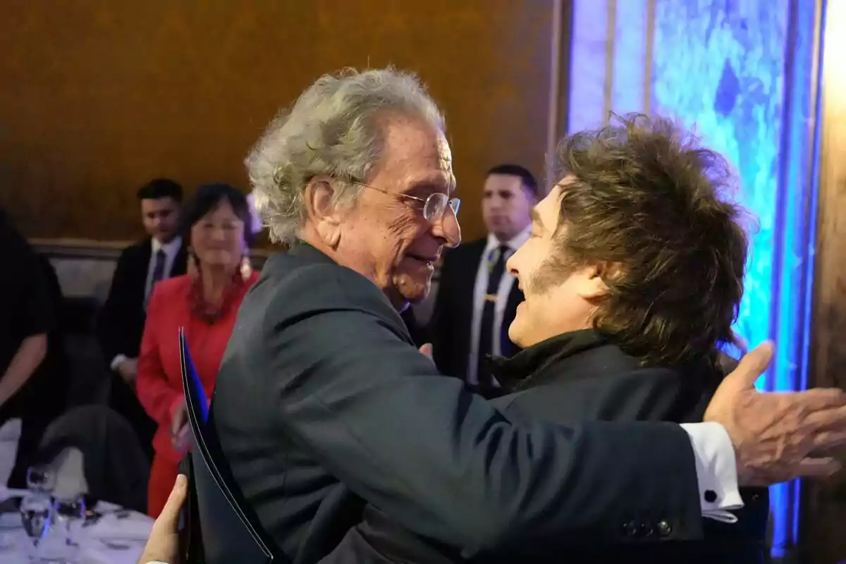 Dos hombres se abrazan sonrientes en un evento formal mientras varias personas observan en el fondo