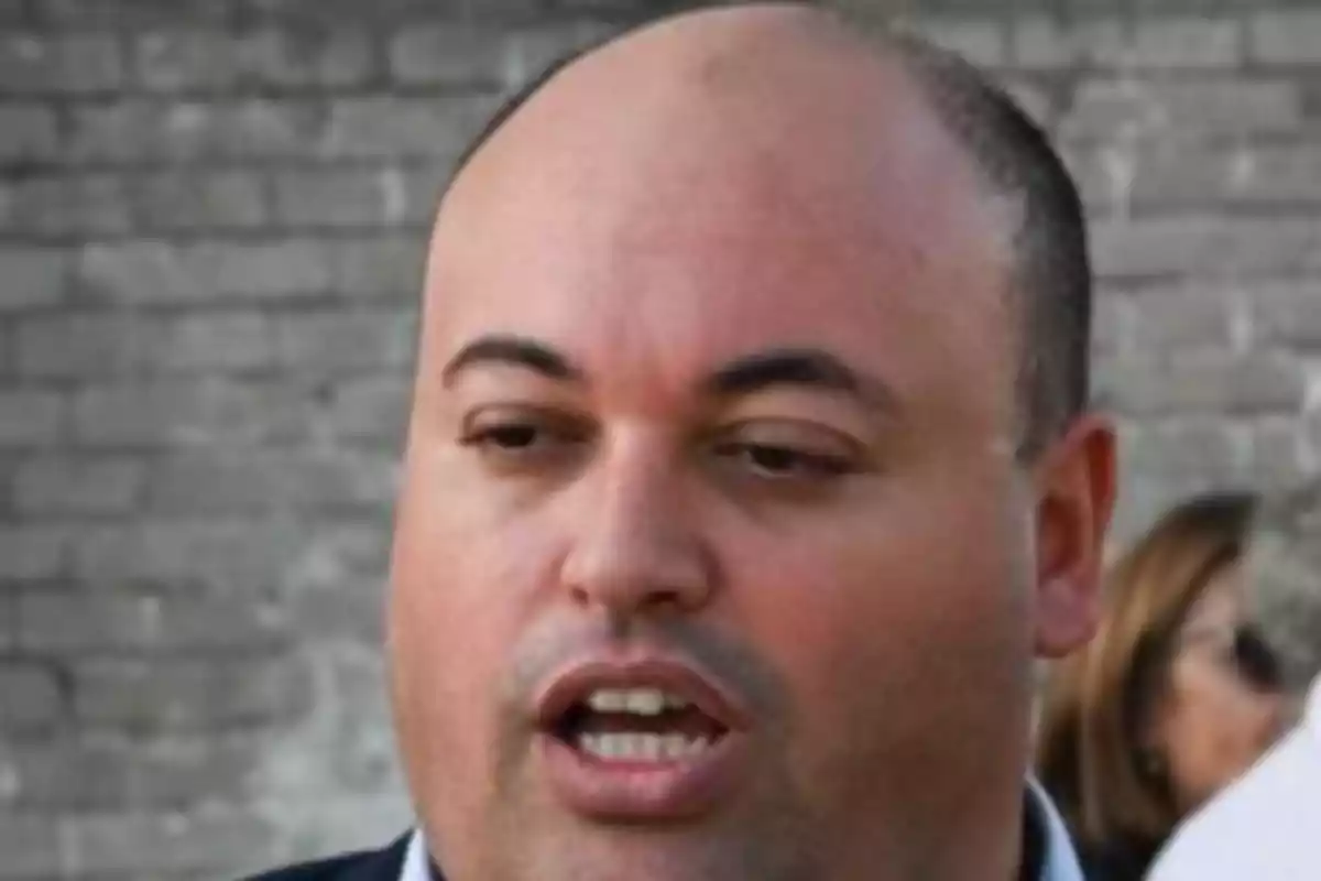 Light-skinned bald man speaking outdoors with a brick wall in the background Light-skinned bald man speaking outdoors with a brick wall in the background