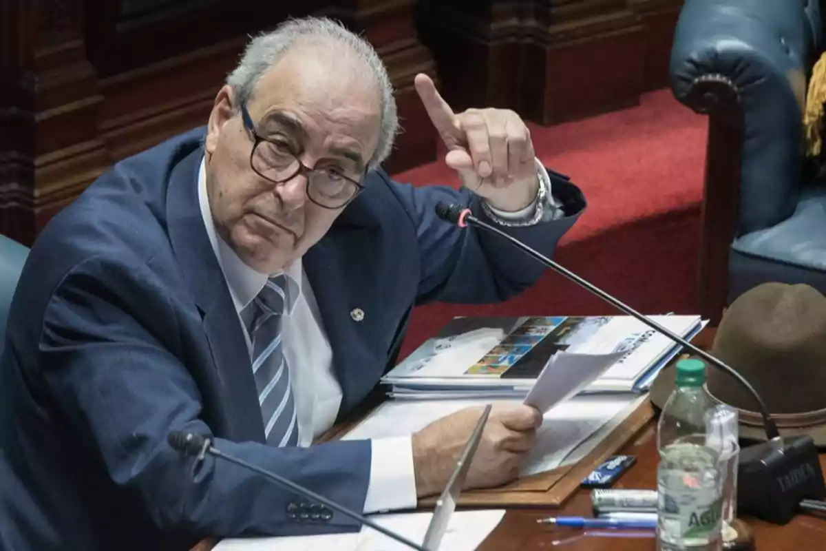 Older man with glasses and a blue suit sitting at a desk raising his hand while holding papers Older man with glasses and a blue suit sitting at a desk raising his hand while holding papers