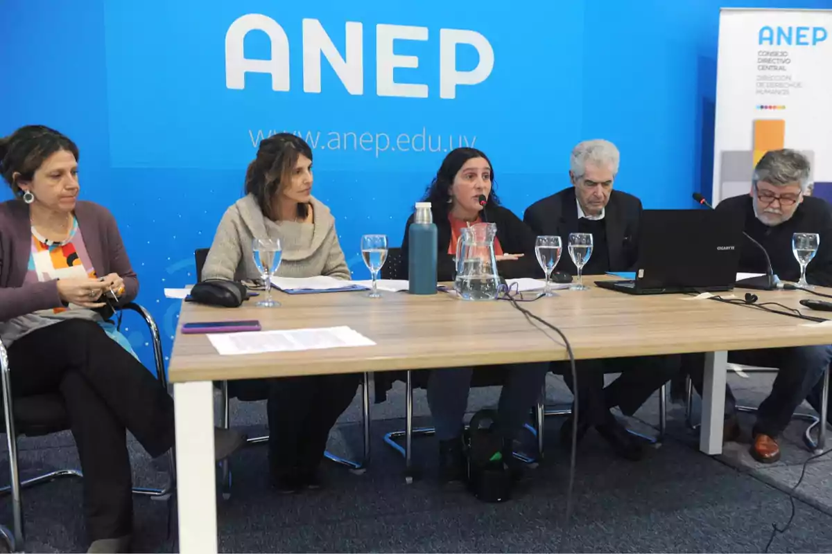 Five people seated at a conference table with microphones, glasses of water, and a laptop, at an ANEP event with a blue background.