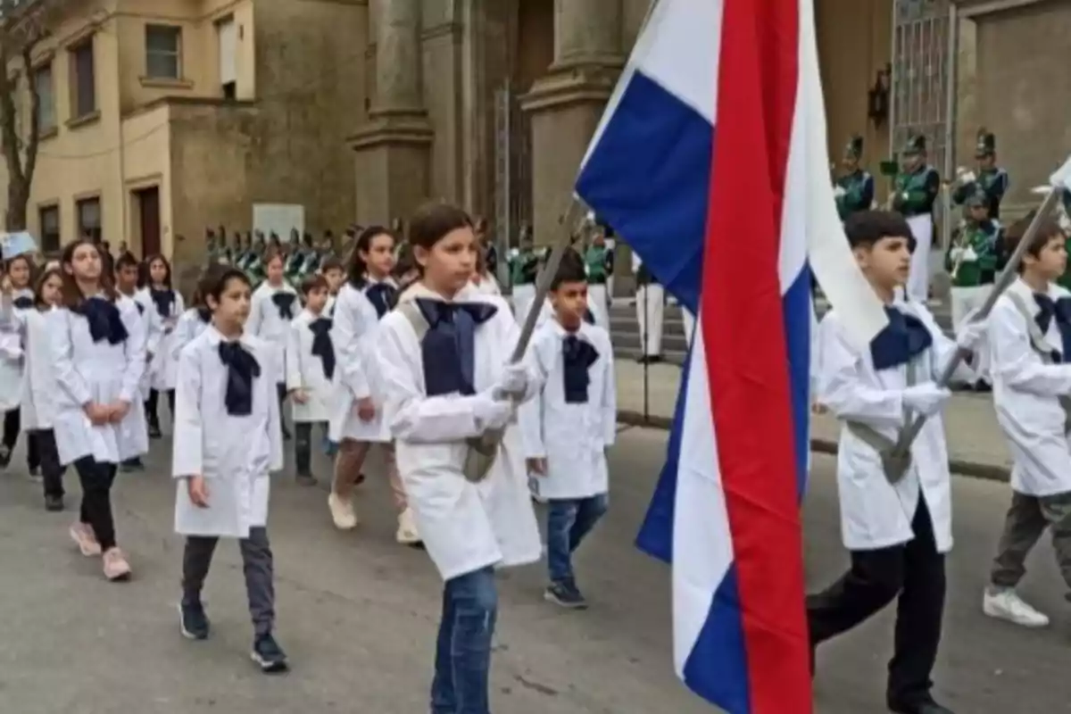 Children in white smocks and blue bows parade down the street carrying a tricolor flag in front of an old building. Children in white smocks and blue bows parade down the street carrying a tricolor flag in front of an old building.