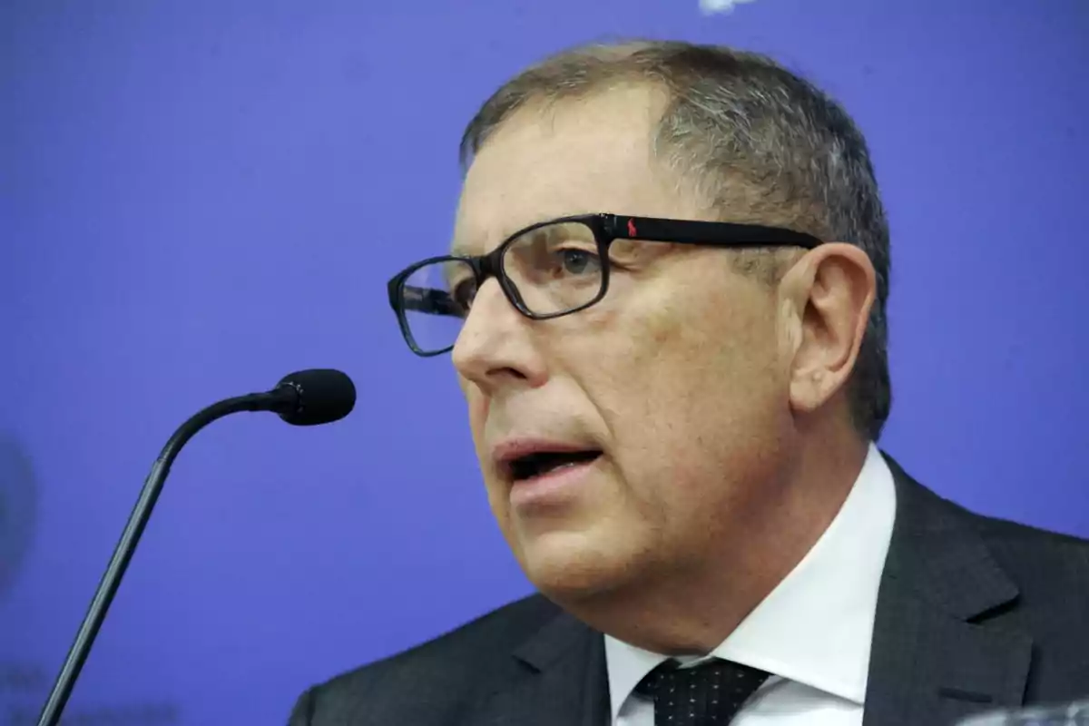 Hombre de mediana edad con gafas y traje hablando frente a un micrófono sobre un fondo azul Hombre de mediana edad con gafas y traje hablando frente a un micrófono sobre un fondo azul