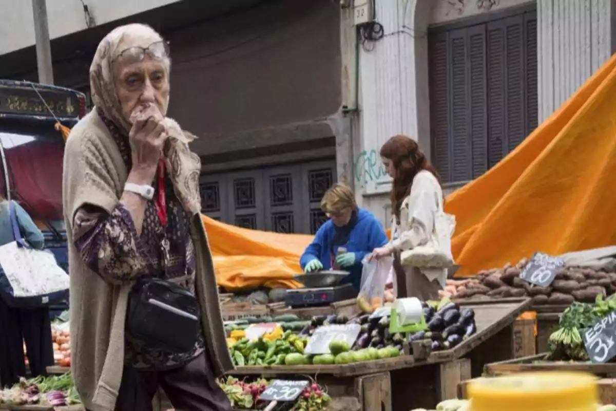 Una mujer mayor con pañuelo en la cabeza camina por un mercado al aire libre donde otras personas compran verduras y hortalizas.
