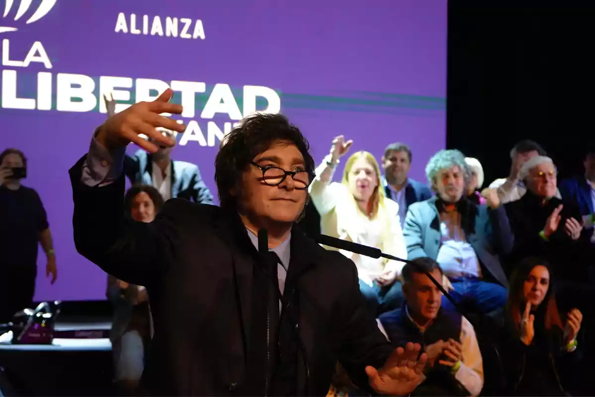 Man with glasses and a dark suit gesturing in front of a microphone, with a group of people applauding and a purple screen with blurry text in the background.