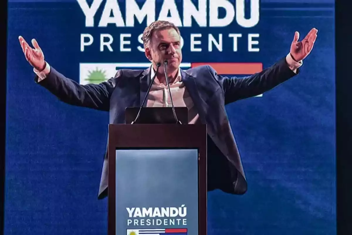 Man in a suit speaking at a podium with his arms outstretched in front of a screen that says Yamandú President