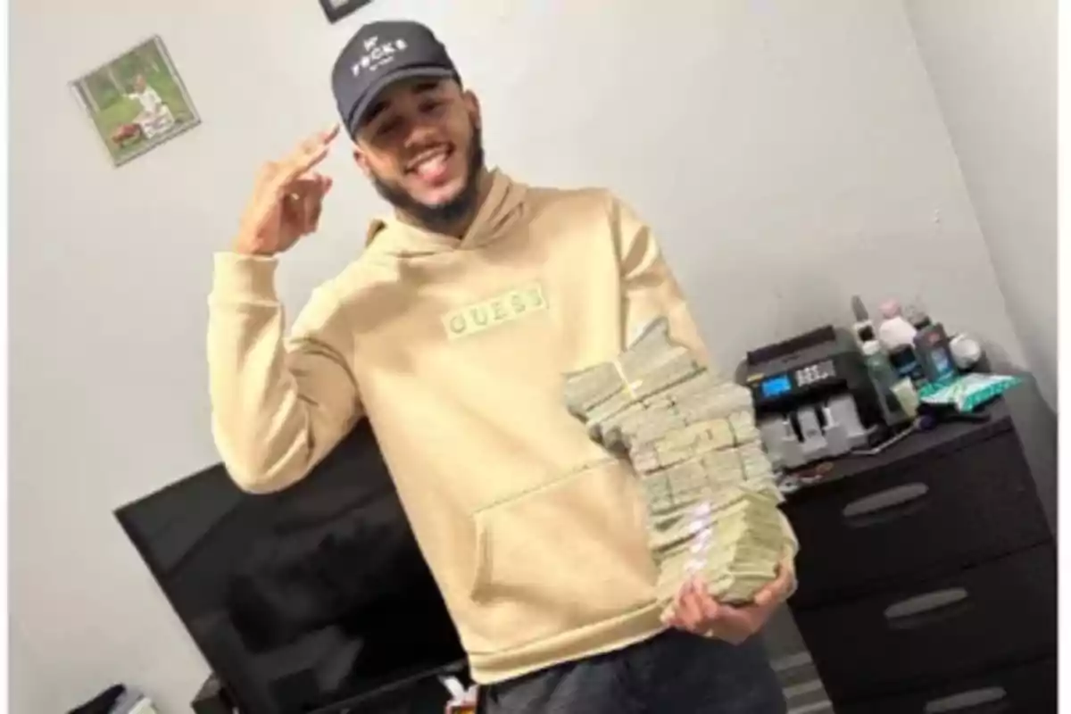 Joven sonriente con barba y gorra, vestido con sudadera beige, sostiene varios fajos de billetes en una habitación con televisor y muebles oscuros.