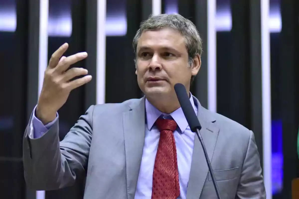 Hombre de traje gris y corbata roja hablando frente a un micrófono con gesto expresivo Hombre de traje gris y corbata roja hablando frente a un micrófono con gesto expresivo