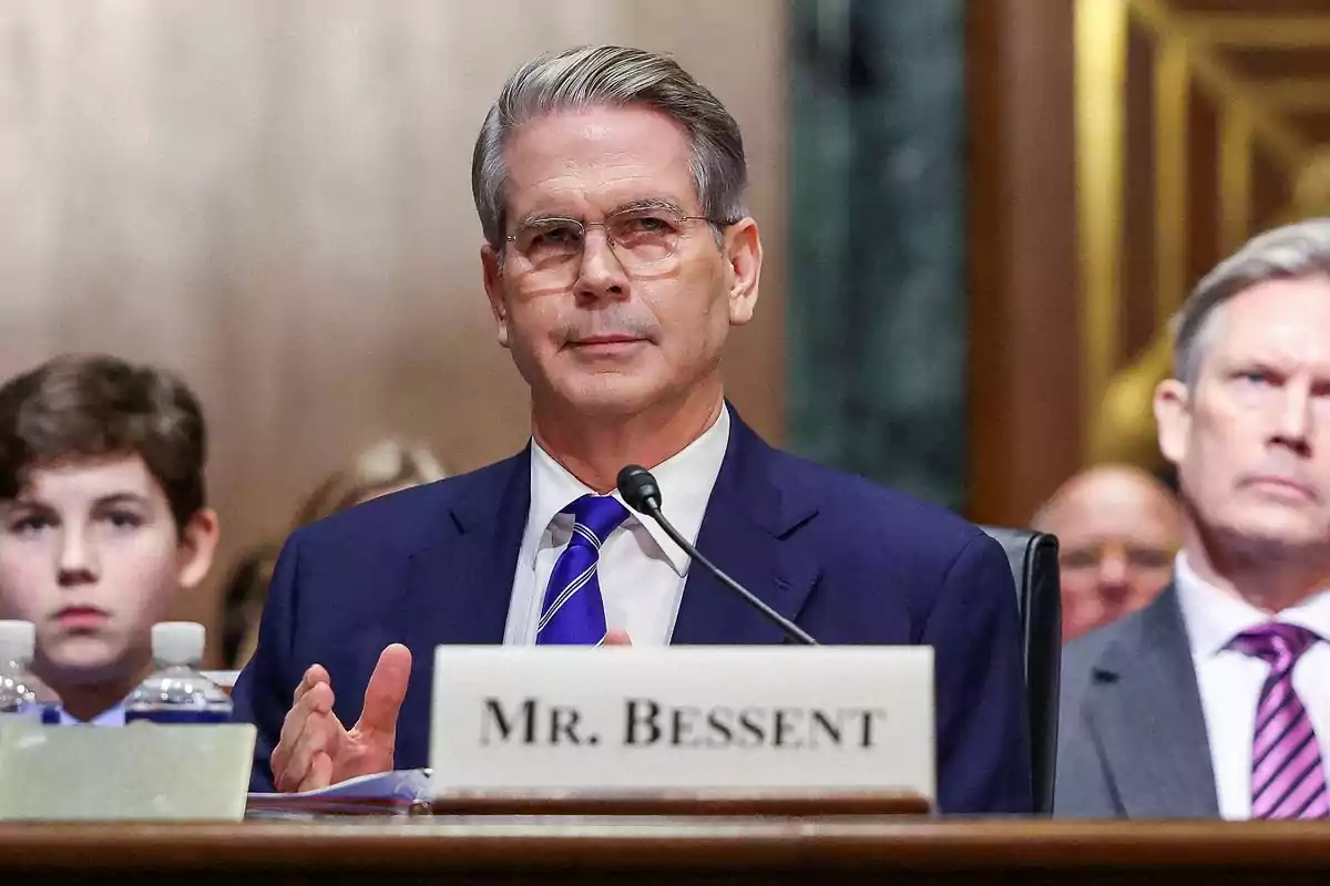 Hombre de cabello canoso y gafas con traje azul sentado frente a un micrófono en una audiencia pública con un letrero que dice MR. BESSENT Hombre de cabello canoso y gafas con traje azul sentado frente a un micrófono en una audiencia pública con un letrero que dice MR. BESSENT