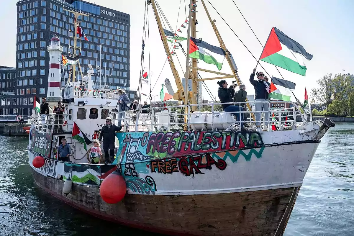 Barco decorado con grafitis y banderas palestinas navegando cerca de un puerto, con varias personas a bordo mostrando pancartas y banderas en apoyo a Palestina, mientras edificios y un faro se ven al fondo.