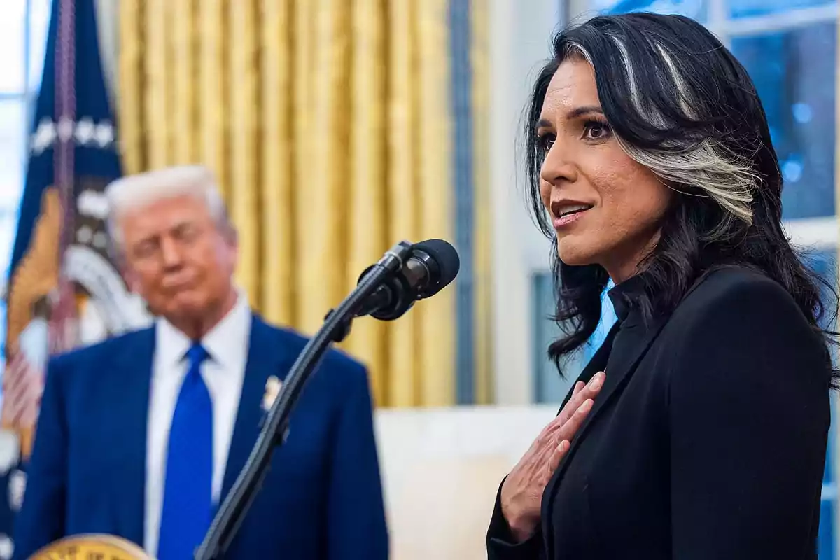 A dark-haired woman with a white streak speaks into a microphone while a blond man in a blue suit watches her from the background in a room with golden curtains.