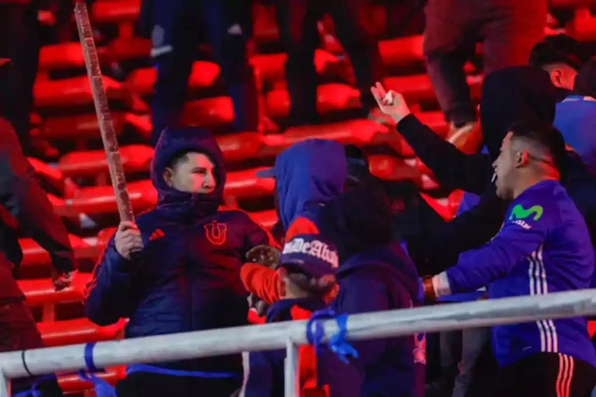 Fans wearing blue clothes argue and one holds a stick in a stand lit with red lights.