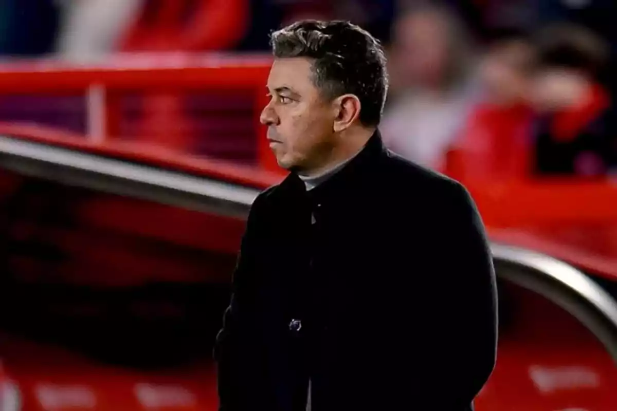Hombre de cabello corto y canoso vistiendo un abrigo oscuro de pie en un estadio de fútbol con fondo desenfocado rojo