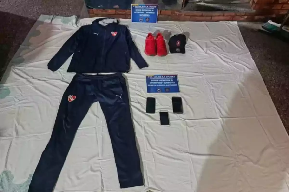 Sobre una sábana blanca hay un conjunto deportivo azul marino con el escudo de un club, un par de zapatillas rojas, una gorra negra, tres teléfonos móviles y dos carteles de la policía de la ciudad Sobre una sábana blanca hay un conjunto deportivo azul marino con el escudo de un club, un par de zapatillas rojas, una gorra negra, tres teléfonos móviles y dos carteles de la policía de la ciudad
