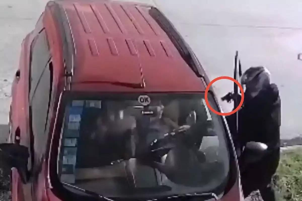 Persona encapuchada sosteniendo un arma junto a la puerta abierta de un automóvil rojo