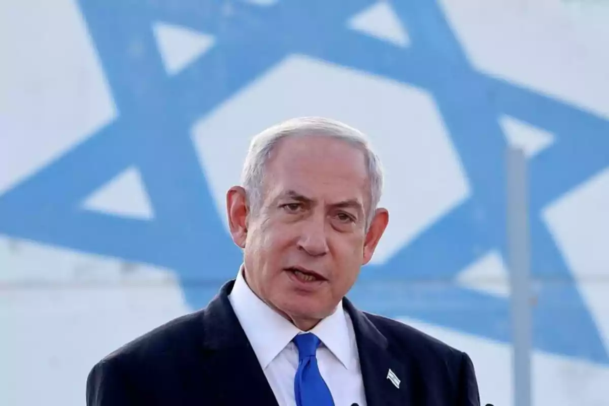 Gray-haired man in a suit and blue tie speaking in front of a blurred Israeli flag Gray-haired man in a suit and blue tie speaking in front of a blurred Israeli flag