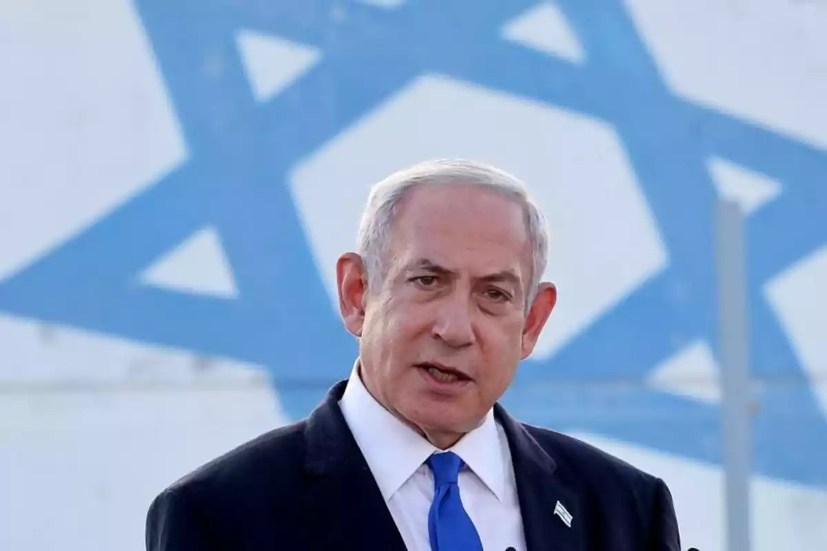 Hombre de cabello canoso con traje y corbata azul hablando frente a una bandera de Israel desenfocada