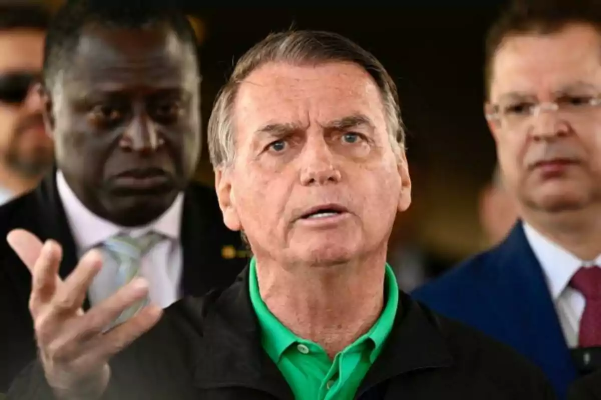 A gray-haired man with a serious expression speaks while gesturing with his hand, accompanied by two people in the background who watch him alertly. A gray-haired man with a serious expression speaks while gesturing with his hand, accompanied by two people in the background who watch him alertly.