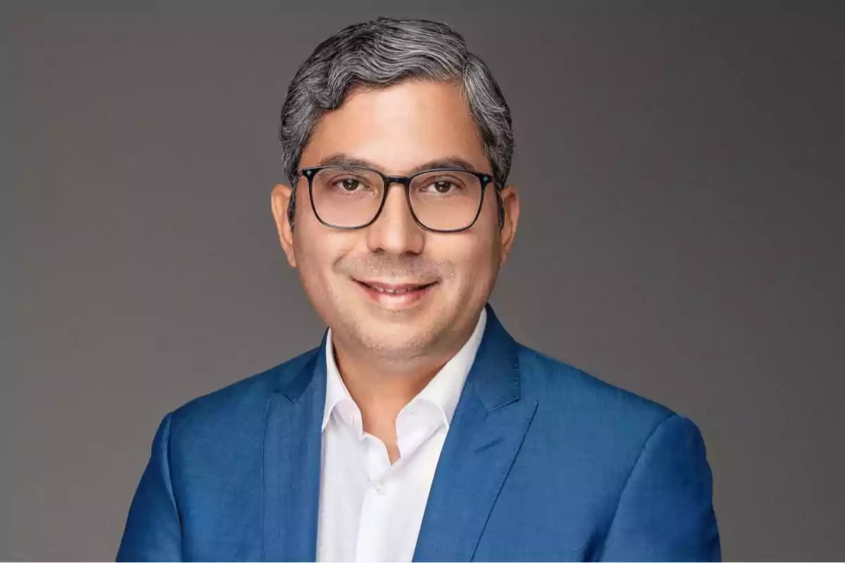 Hombre de cabello canoso y corto con gafas, usando saco azul y camisa blanca, posando frente a un fondo gris Hombre de cabello canoso y corto con gafas, usando saco azul y camisa blanca, posando frente a un fondo gris