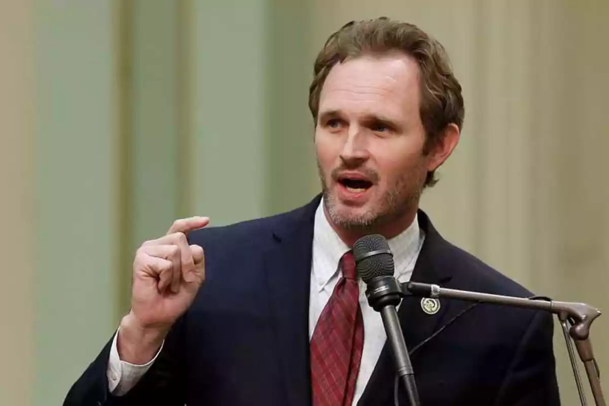 Brown-haired man with a short beard speaking into a microphone while gesturing with his hand in a formal setting