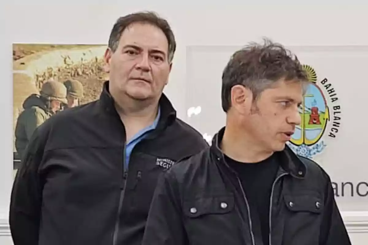 Dos hombres de pie en un ambiente interior, uno mirando al frente y el otro de perfil, con un escudo de Bahía Blanca en la pared detrás de ellos. Dos hombres de pie en un ambiente interior, uno mirando al frente y el otro de perfil, con un escudo de Bahía Blanca en la pared detrás de ellos.