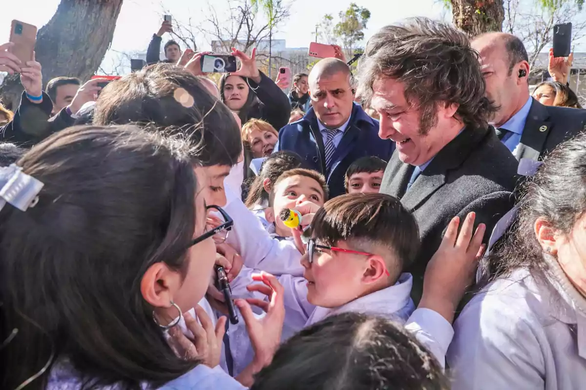 Un hombre sonriente rodeado de niños con guardapolvos blancos mientras varias personas toman fotos con sus teléfonos en un ambiente al aire libre. Un hombre sonriente rodeado de niños con guardapolvos blancos mientras varias personas toman fotos con sus teléfonos en un ambiente al aire libre.