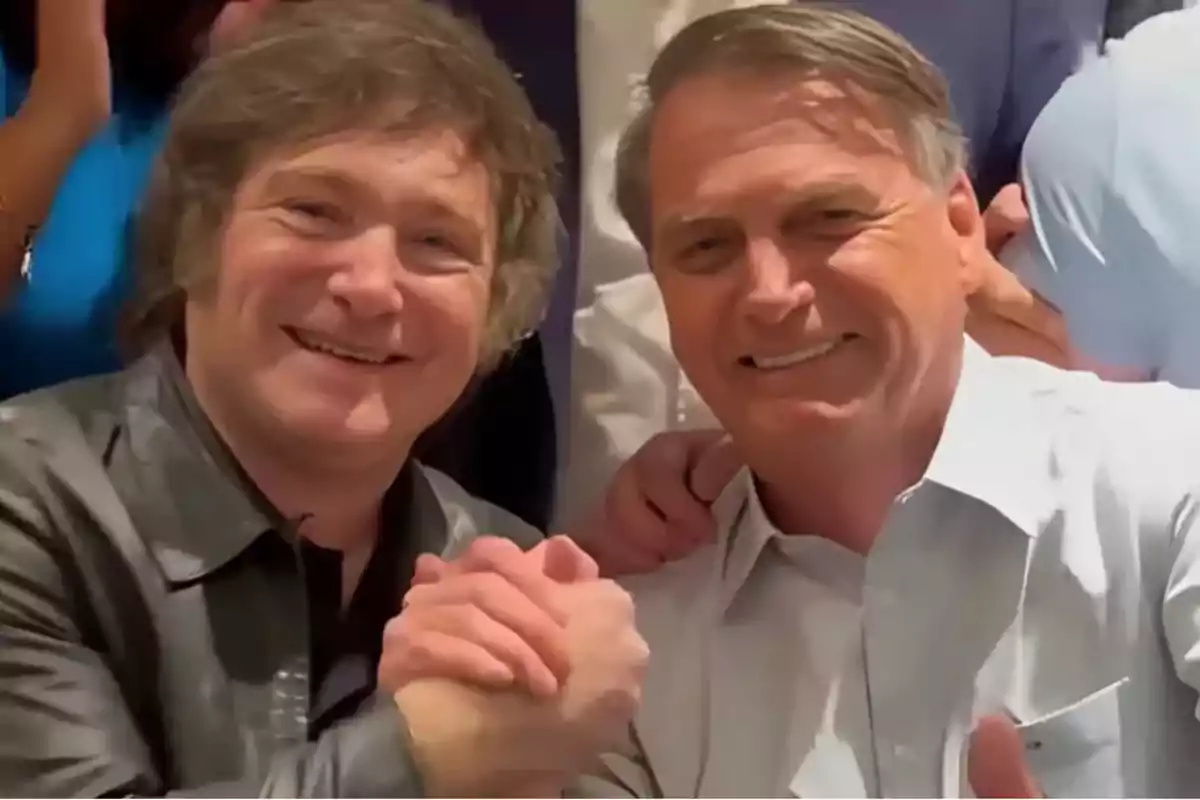 Two men smile and shake hands in a show of camaraderie while looking at the camera.