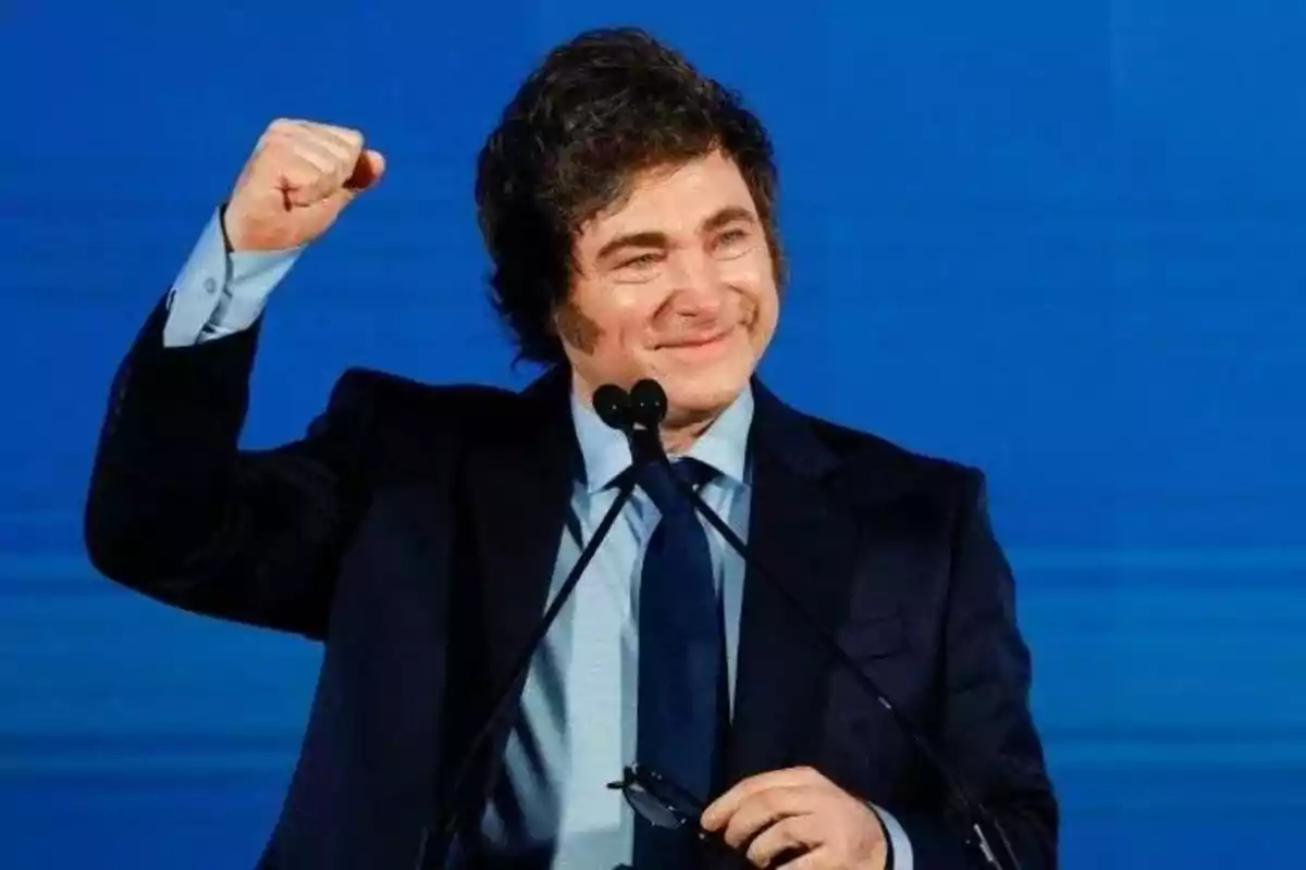 Hombre de cabello oscuro con traje y corbata levantando el puño y sonriendo frente a un micrófono y un fondo azul