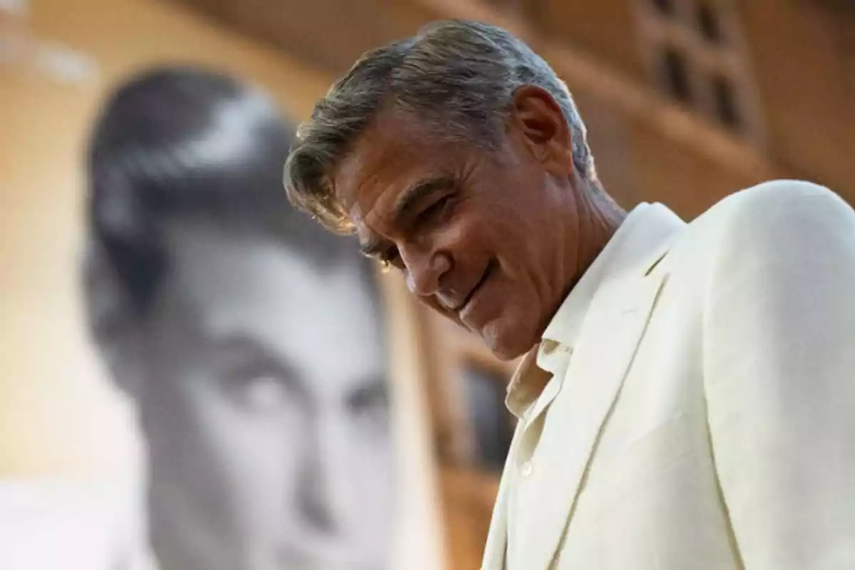 Hombre de cabello canoso con saco blanco sonriendo mientras mira hacia abajo, con un retrato desenfocado en el fondo Hombre de cabello canoso con saco blanco sonriendo mientras mira hacia abajo, con un retrato desenfocado en el fondo