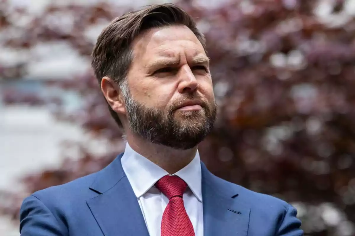 Hombre de barba y cabello corto con traje azul y corbata roja mirando hacia el horizonte con expresión seria
