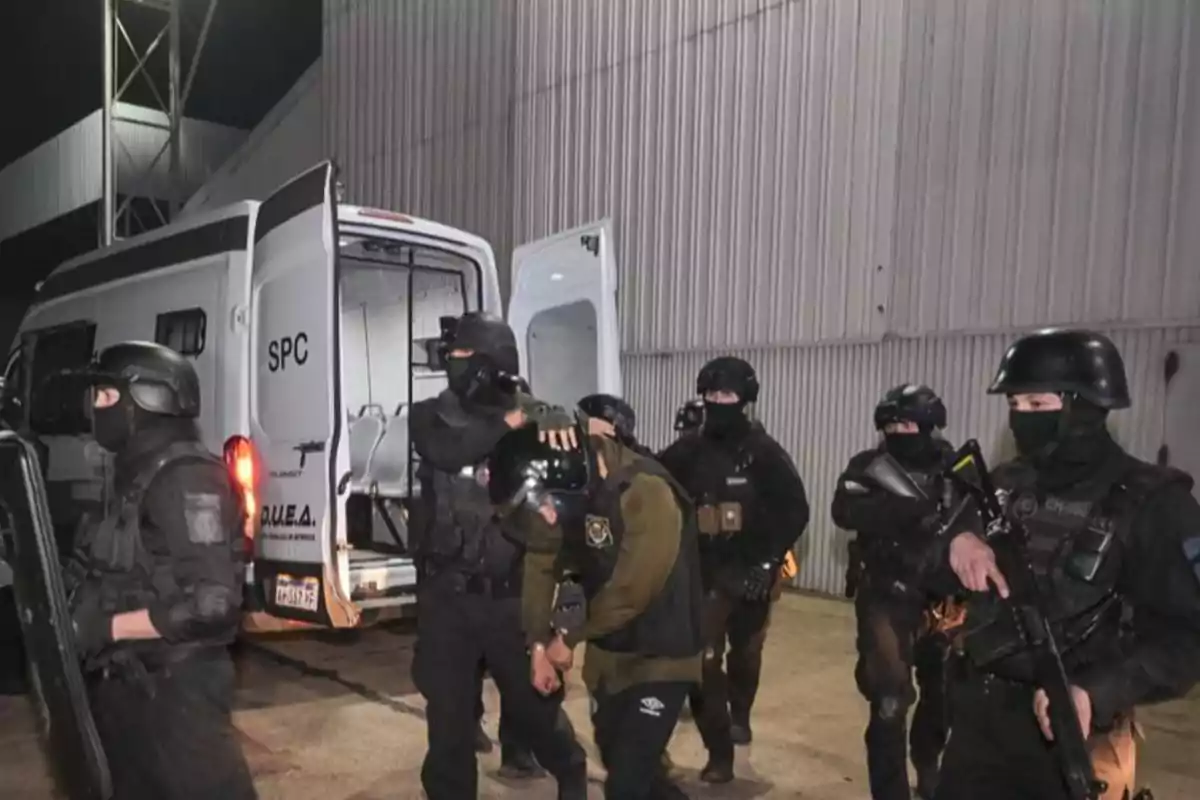 Varios policías fuertemente armados y con equipo táctico escoltan a una persona esposada hacia una camioneta policial durante la noche frente a un edificio industrial.