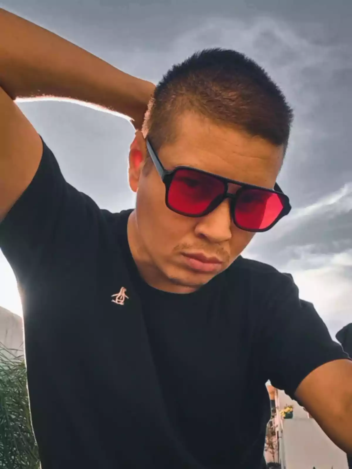 Persona con gafas de sol rojas y camiseta negra posando al aire libre con el brazo detrás de la cabeza y el cielo nublado de fondo Persona con gafas de sol rojas y camiseta negra posando al aire libre con el brazo detrás de la cabeza y el cielo nublado de fondo