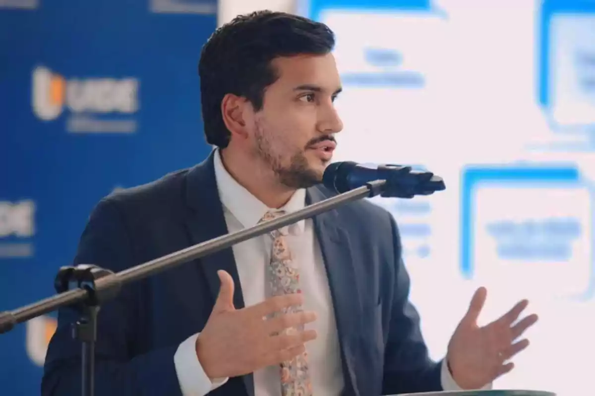 Hombre joven con barba usando traje y corbata hablando frente a un micrófono en un evento profesional con fondo azul desenfocado