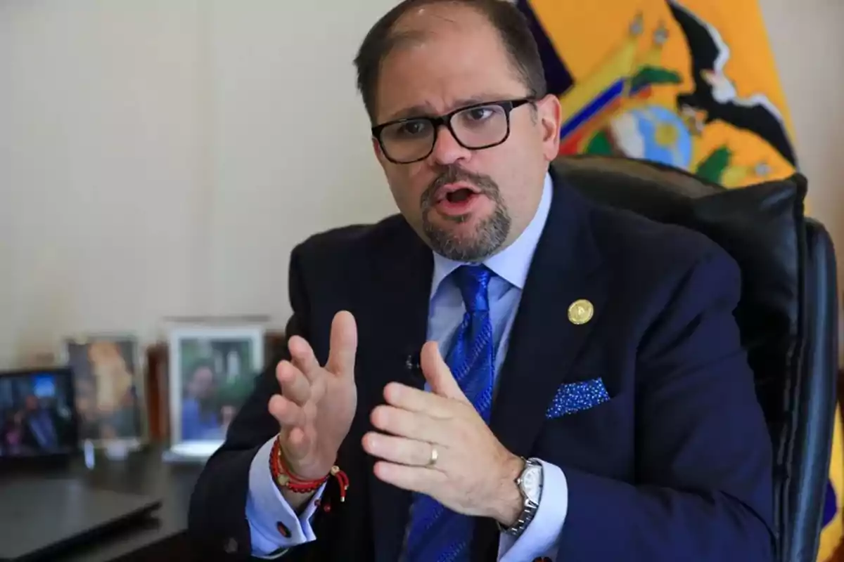 Hombre con gafas y traje azul hablando con gestos sentado en una oficina con la bandera de Ecuador al fondo