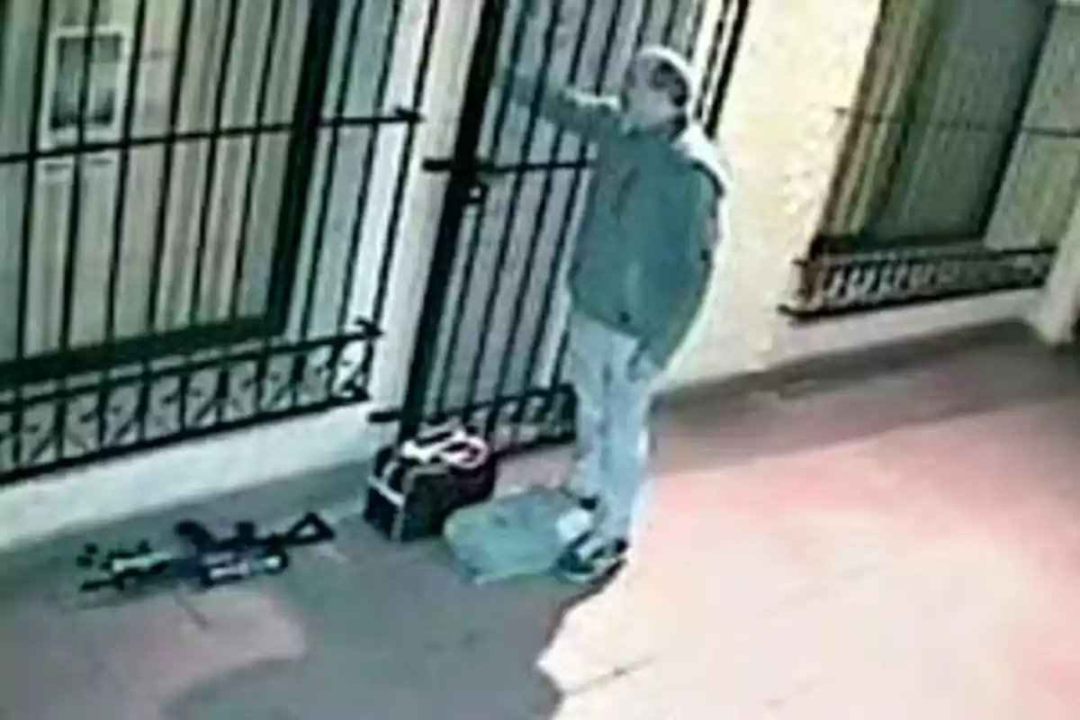 Hombre de pie frente a una reja en una zona iluminada con varios objetos en el suelo junto a él Hombre de pie frente a una reja en una zona iluminada con varios objetos en el suelo junto a él