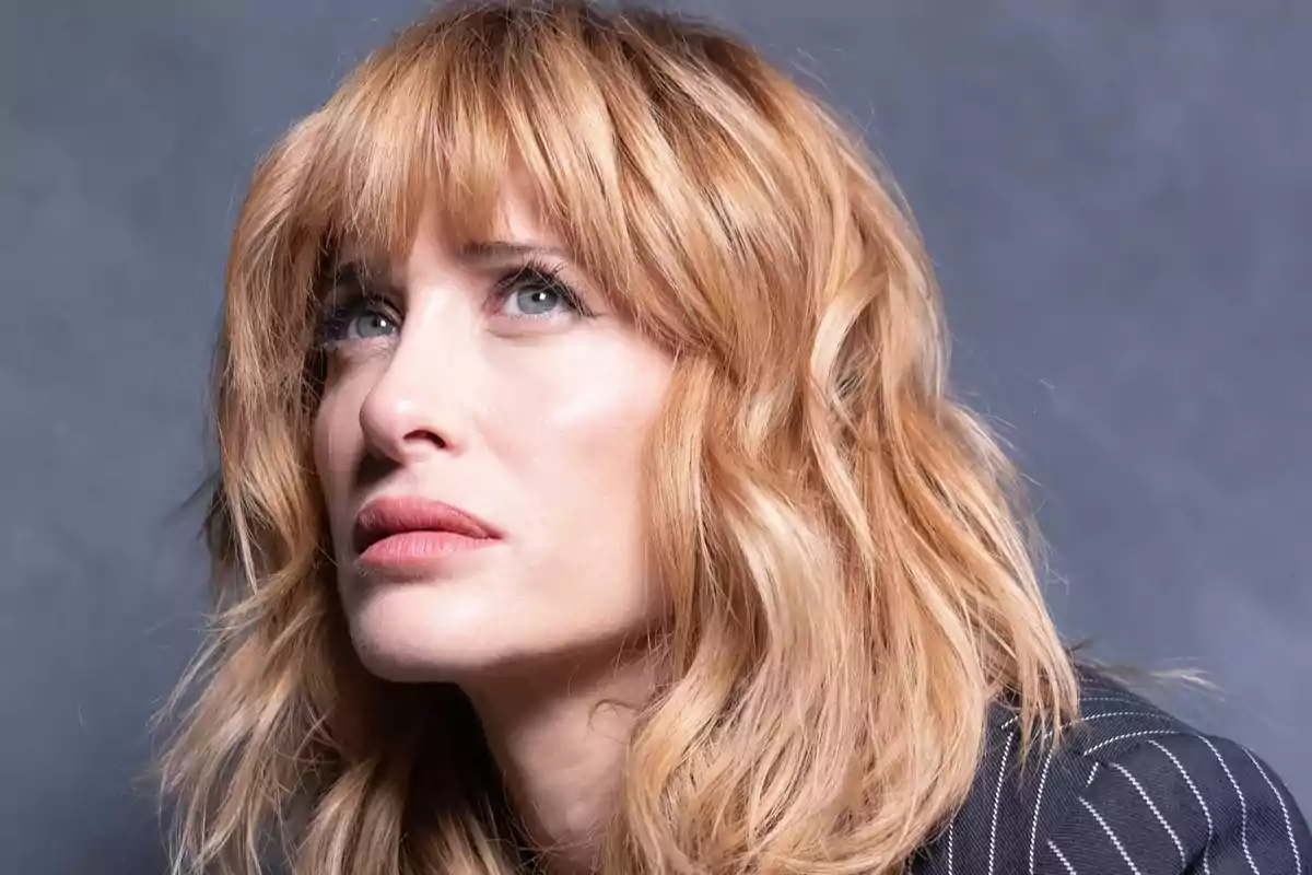 Mujer de cabello rubio ondulado y ojos claros mirando hacia arriba con expresión seria sobre fondo gris