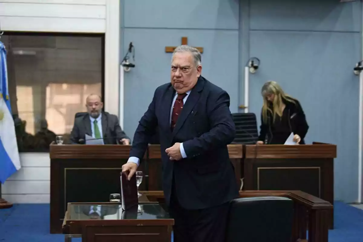 Man in a suit in a courtroom with a serious expression while holding a book; in the background, there are two people seated and an Argentine flag.