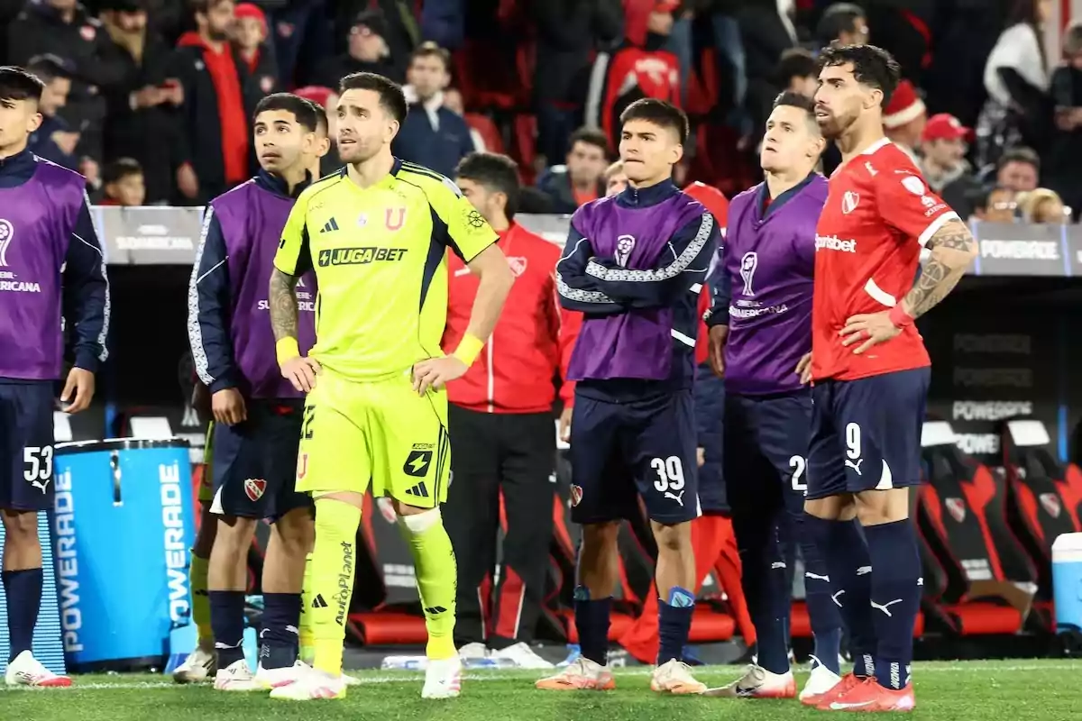 Soccer players from two teams watch alertly on the field during a match, some wearing uniforms and others wearing substitute bibs, while the crowd can be seen in the stands in the background. Soccer players from two teams watch alertly on the field during a match, some wearing uniforms and others wearing substitute bibs, while the crowd can be seen in the stands in the background.