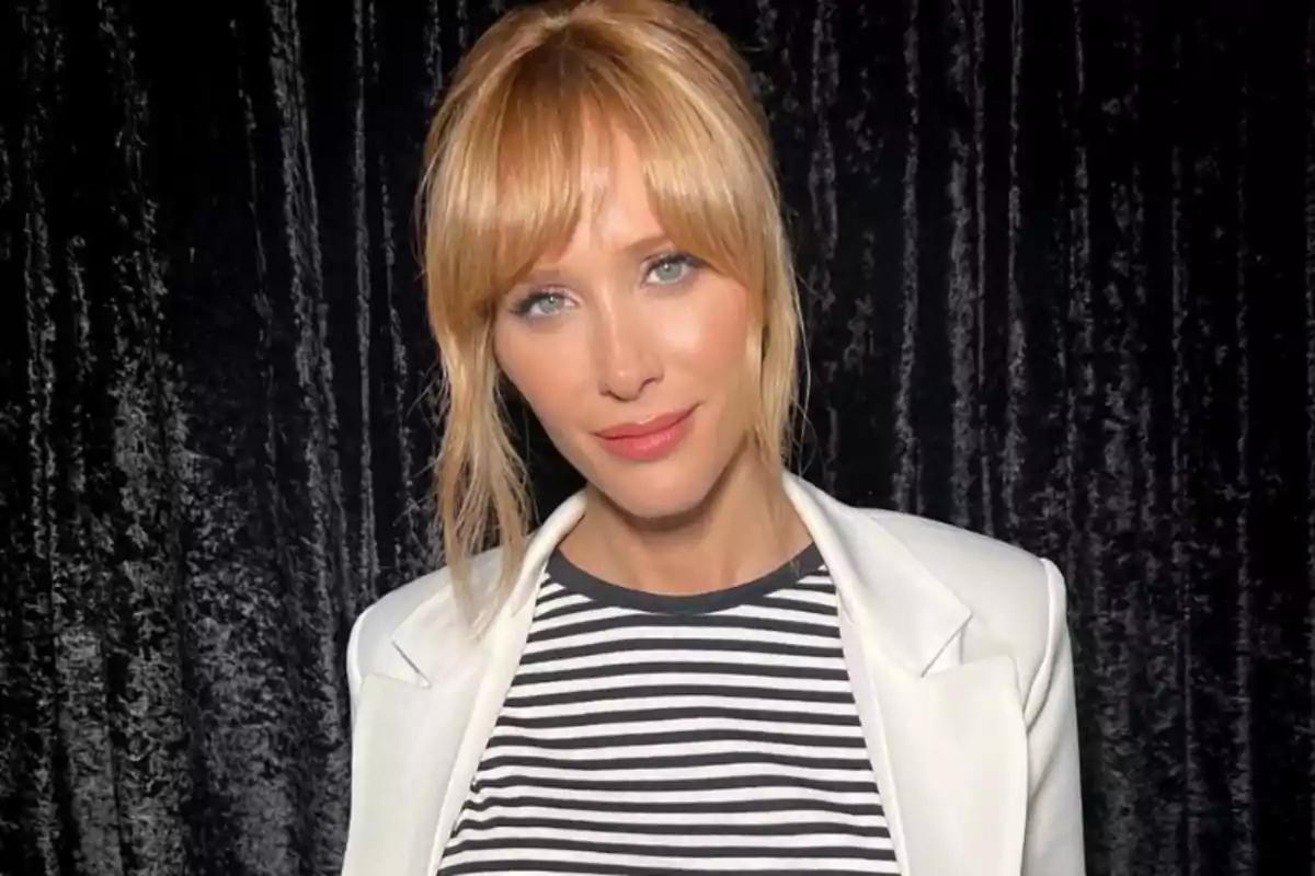 Mujer rubia con flequillo usando saco blanco y camiseta de rayas frente a una cortina negra