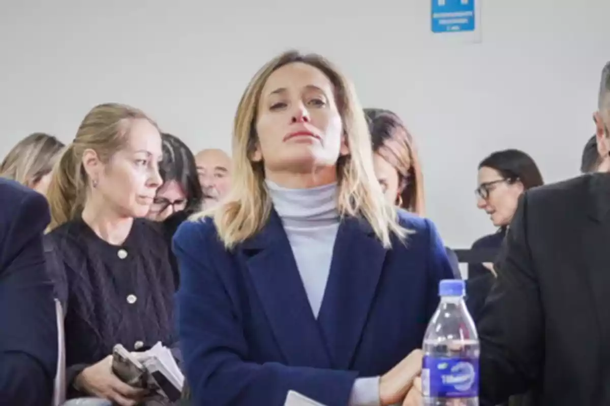 Mujer rubia con expresión seria sentada en una sala junto a otras personas