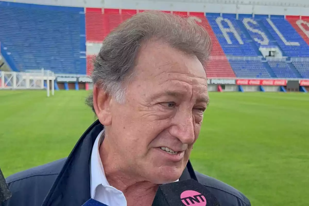 Older man giving an interview in a soccer stadium with red and blue stands in the background