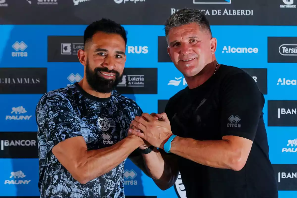Dos hombres sonríen y se saludan con un apretón de manos frente a un fondo con logotipos de patrocinadores deportivos Dos hombres sonríen y se saludan con un apretón de manos frente a un fondo con logotipos de patrocinadores deportivos