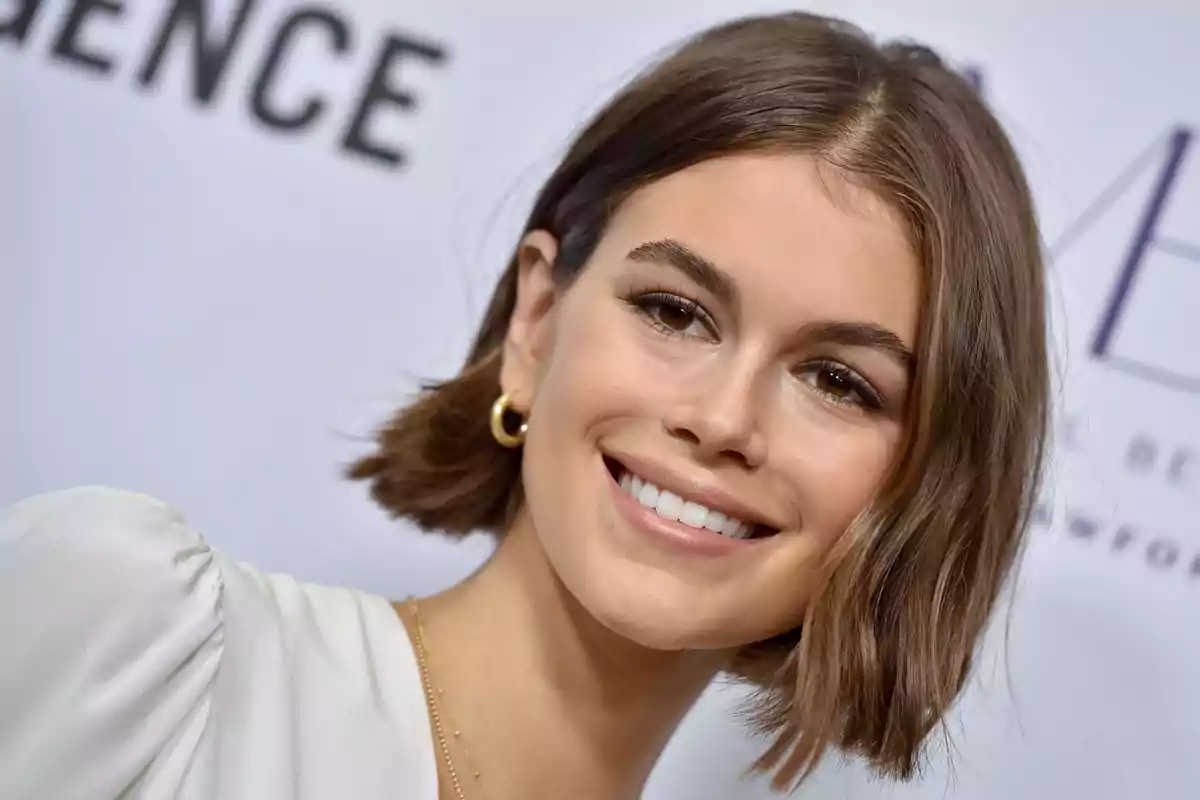 Joven con cabello corto castaño y sonrisa amplia usando aretes dorados y blusa blanca en un evento Joven con cabello corto castaño y sonrisa amplia usando aretes dorados y blusa blanca en un evento