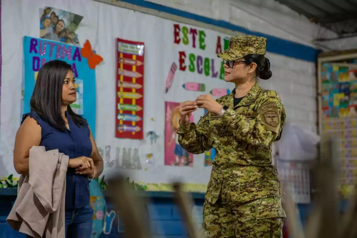 Una mujer vestida de civil conversa con una mujer militar en un aula decorada con carteles coloridos y mensajes escolares. Una mujer vestida de civil conversa con una mujer militar en un aula decorada con carteles coloridos y mensajes escolares.