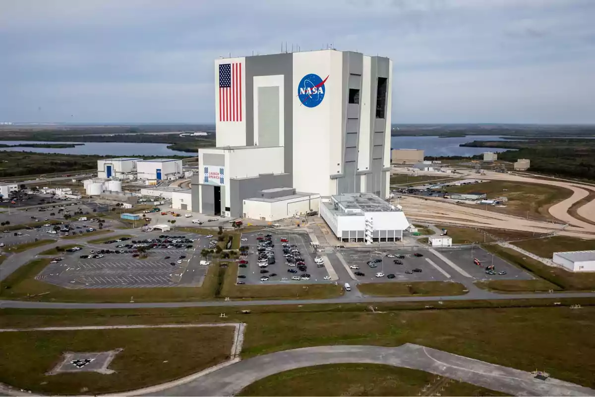Edificio grande de la NASA con una bandera de Estados Unidos y un estacionamiento lleno de autos rodeado de áreas verdes y cuerpos de agua Edificio grande de la NASA con una bandera de Estados Unidos y un estacionamiento lleno de autos rodeado de áreas verdes y cuerpos de agua