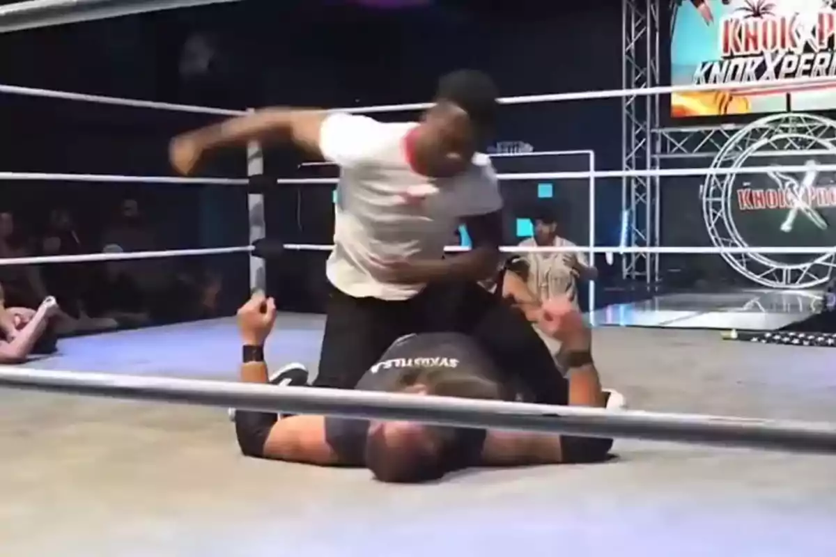 Dos luchadores están en el ring, uno de ellos está en el suelo mientras el otro lo golpea con el puño, alrededor se observa público y un letrero de evento de lucha libre