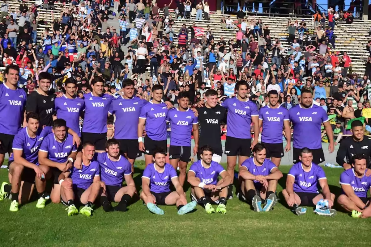 Rugby team posing for a group photo on the field wearing purple jerseys with a crowd of fans in the stands in the background Rugby team posing for a group photo on the field wearing purple jerseys with a crowd of fans in the stands in the background